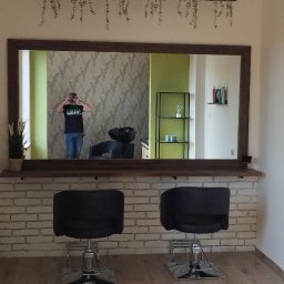 Szymon Szymański - Wnętrze salonu fryzjerskiego z dwoma czarnymi fotelami, dużym lustrem z drewnianą ramą, oświetleniem punktowym i roślinami wiszącymi nad lustrem, widok na ścianę z cegły i podłogę z paneli.