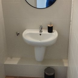 Szymon Szymański - Umywalka ceramiczna zamontowana na ścianie w łazience z brązowymi płytkami podłogowymi i okrągłym lustrem z jutowym sznurem.