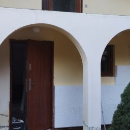 Szymon Szymański - Ocieplanie elewacji budynku z charakterystycznymi łukami, widoczne płyty styropianowe przed położeniem warstwy wykończeniowej, częściowo odsłonięte ocieplenie przy oknie.