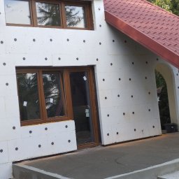 Szymon Szymański - Ocieplanie elewacji styropianem w Gajkowicach, widoczne okna z brązowymi ramami i czerwony dach, elewacja przygotowana do nałożenia tynku.