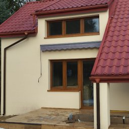 Szymon Szymański - Jasna elewacja domu z brązowymi oknami i czerwonym dachem, widoczne drewniane stopnie tarasu i resztki śniegu na ziemi.
