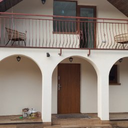 Szymon Szymański - Dwukondygnacyjny dom z kremową elewacją, balkonem z metalową balustradą i trzema łukowatymi wejściami, otoczony drzewami.