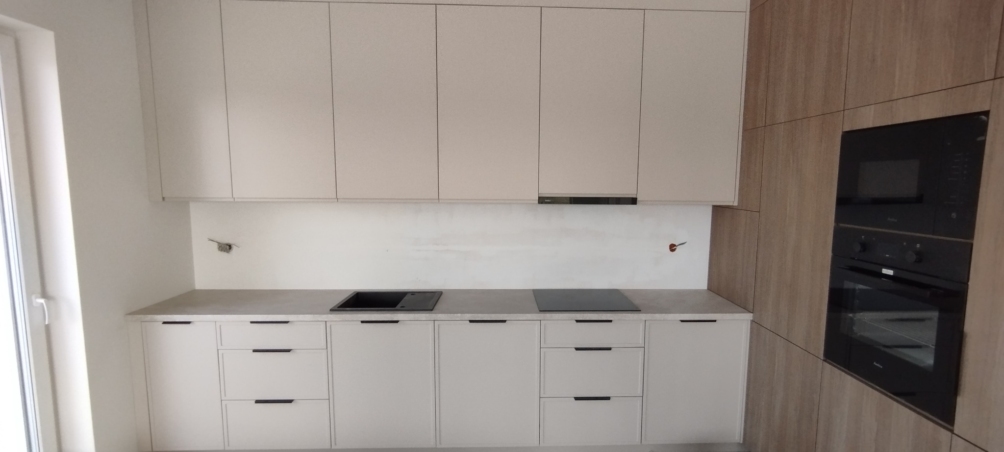 Nowoczesna kuchnia z beżowymi szafkami i drewnianą zabudową piekarnika, widoczne gniazdka elektryczne czekające na podłączenie sprzętu, minimalistyczny design.