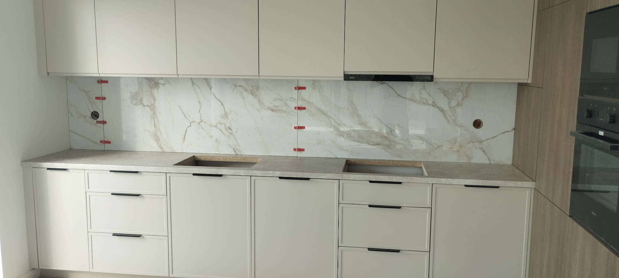 Montaż marmurowej płyty nad blatem kuchennym, z widocznymi czerwonymi krzyżykami dystansowymi i otworami na gniazdka, podkreślający etap prac wykończeniowych.