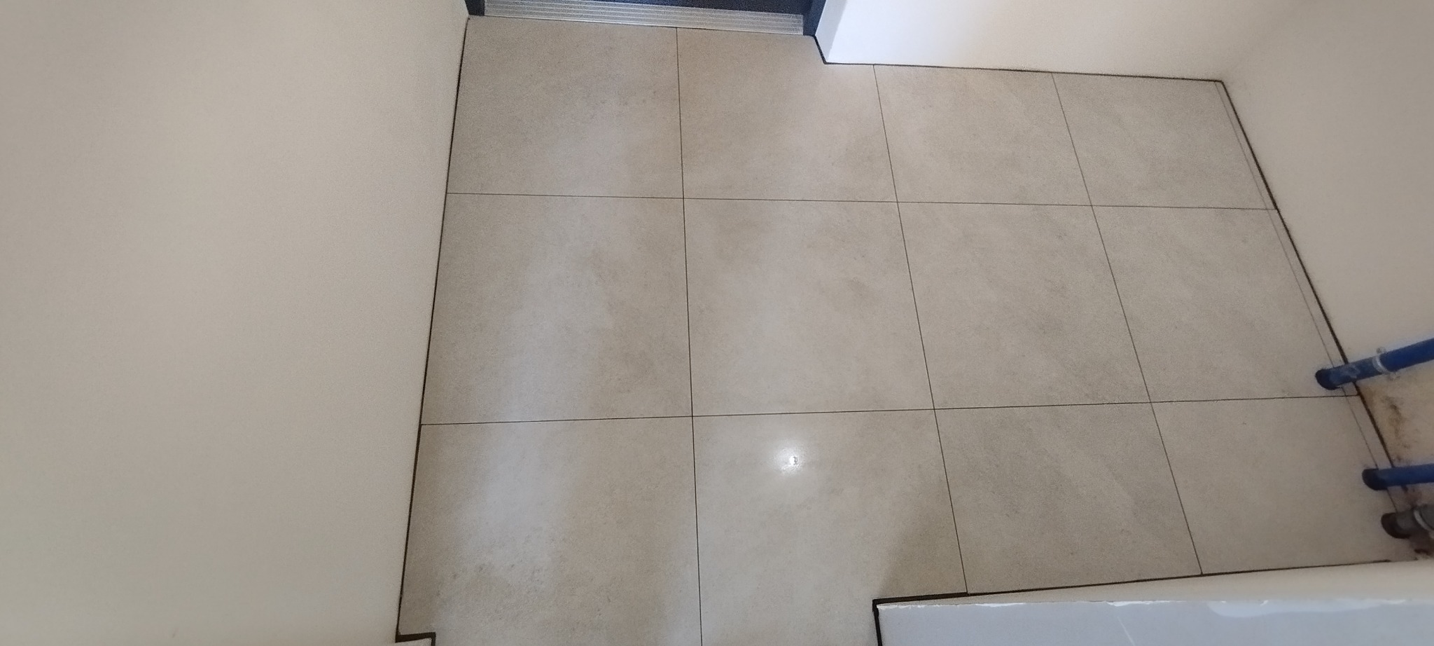 Beżowe płytki podłogowe ułożone w kwadraty, widoczne fugi, fragment ściany i rury. Układanie płytek w małej łazience. Minimalistyczny styl wykończenia.