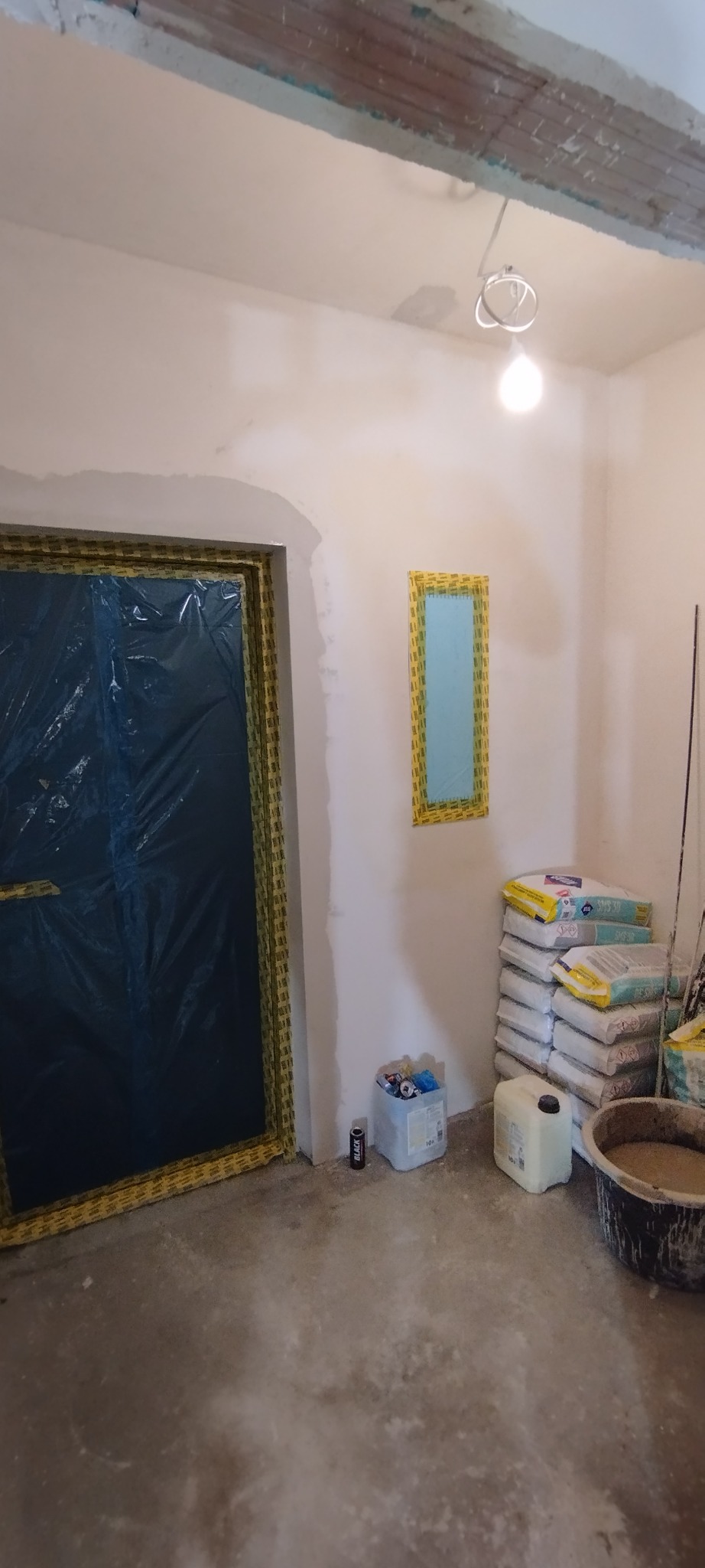 Pokój w trakcie remontu: zabezpieczone folią drzwi i okno, worki z gładzią, wiadro z zaprawą. Widoczne ślady gładzi na ścianach. Oświetlenie żarówką.