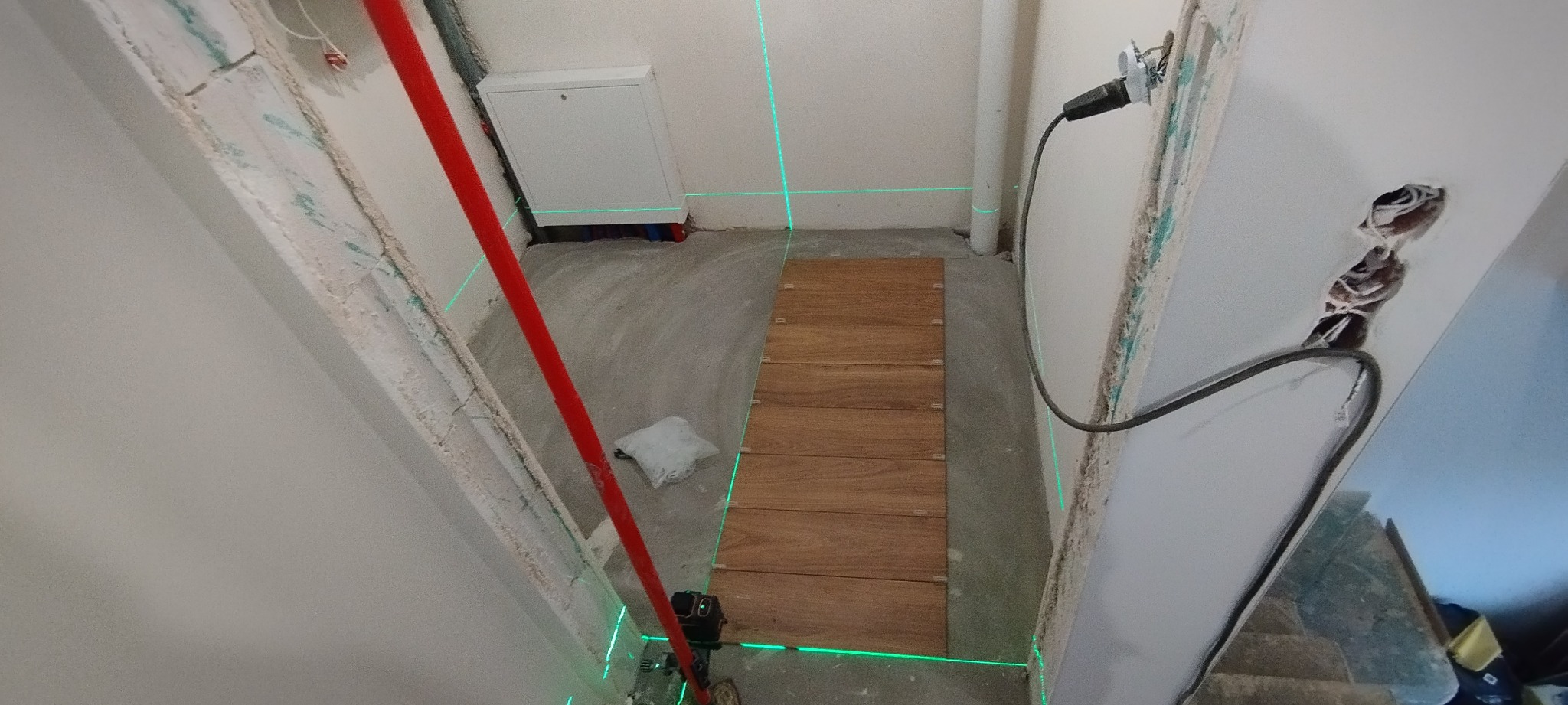 Fragment układania paneli podłogowych w korytarzu w Gajkowicach, z użyciem lasera krzyżowego do precyzyjnego poziomowania. Widoczne przygotowane podłoże i surowe ściany.
