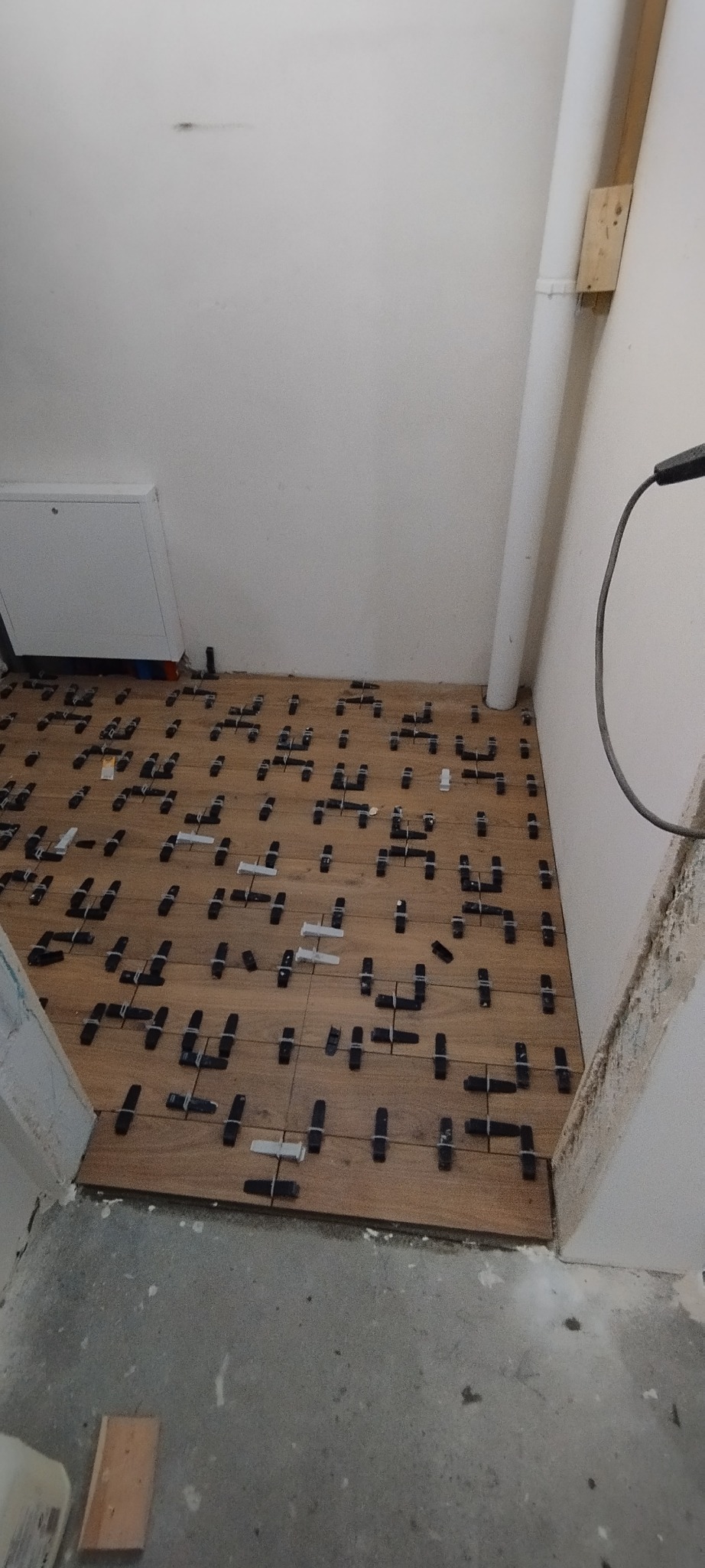 Układanie drewnopodobnych płytek podłogowych z użyciem klipsów poziomujących w narożnym pomieszczeniu. Widoczny fragment rury przy ścianie.