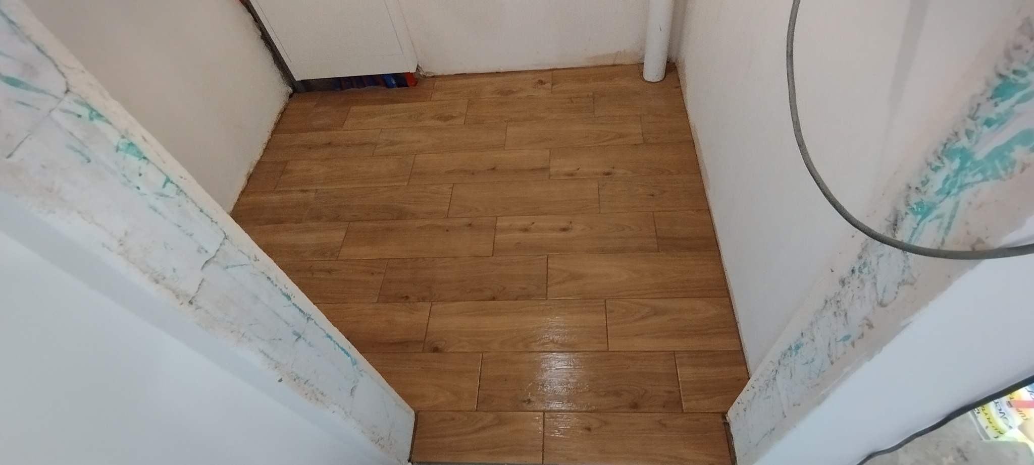Ułożone płytki podłogowe imitujące drewno w wąskim pomieszczeniu z białymi ścianami. Widoczny fragment rury i niebieskie ślady na framudze.