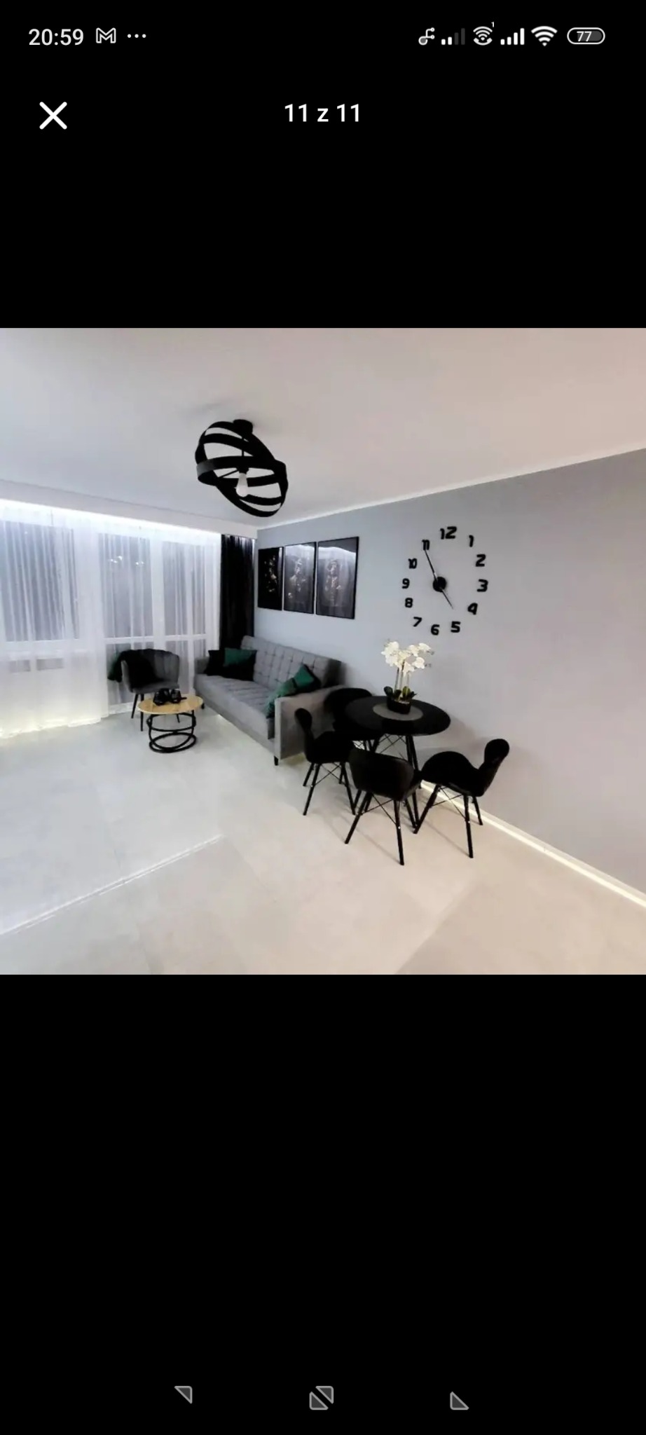 Jasne wnętrze pokoju dziennego: szare ściany, kremowa podłoga, szara sofa, czarne krzesła i stół. Na ścianie duży zegar. Nowoczesne oświetlenie i dekoracje.