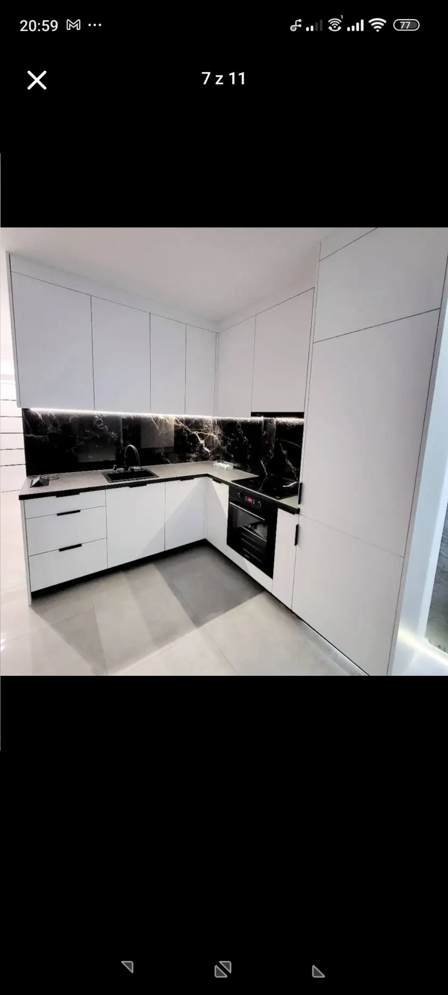 Biała, narożna kuchnia z czarnym blatem i marmurowym panelem nad blatem. Nowoczesny design z minimalistycznymi uchwytami i oświetleniem LED.