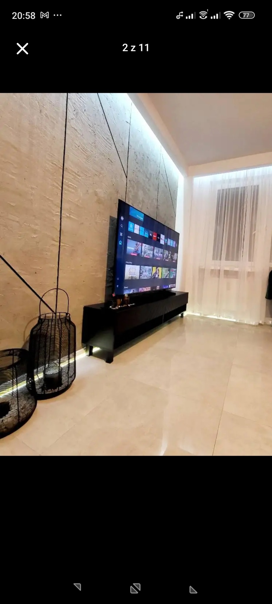 Nowoczesny salon z telewizorem na czarnej szafce RTV, dekoracyjną ścianą w beżowych tonacjach i dużym oknem z białymi zasłonami. Uwagę przyciągają detale dekoracyjne.