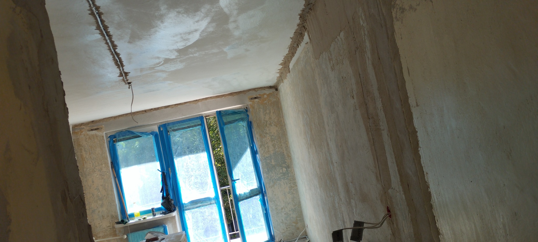 Pokój w trakcie prac wykończeniowych: ściany przygotowane do malowania, okna zabezpieczone folią. Widoczne nierówności ścian i sufitu, instalacja elektryczna.