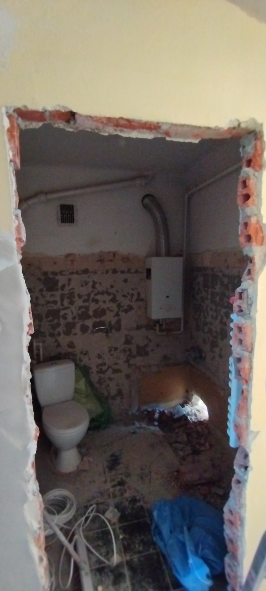 Wnętrze łazienki w trakcie remontu: widoczne stare płytki, toaleta, instalacja grzewcza oraz gruz. Ściany częściowo usunięte, odsłonięta cegła.