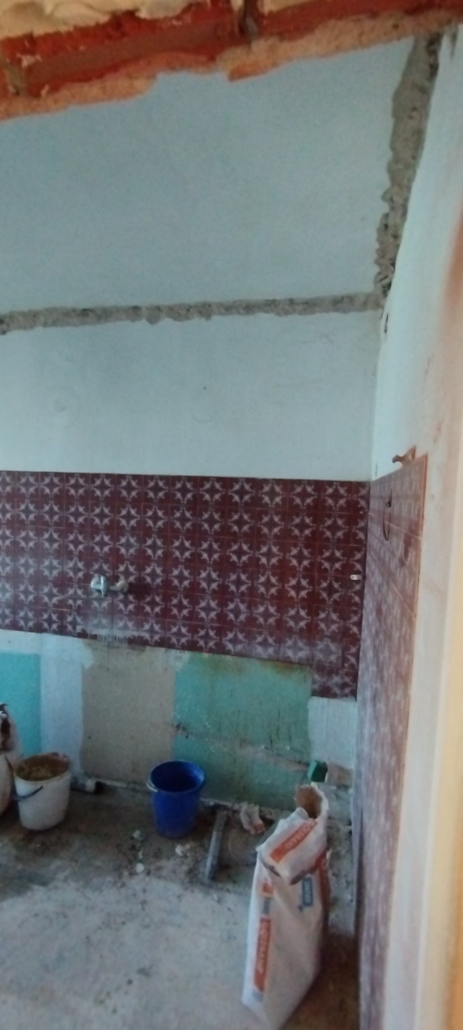 Wnętrze łazienki w trakcie remontu: częściowo zdjęte płytki, widoczne ślady po kleju, wiadro, worek z zaprawą murarską. Przygotowanie do dalszych prac wykończeniowych.