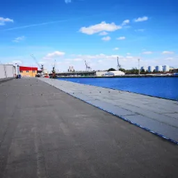 Widok na płaski dach pokryty papą na tle portu z dźwigami i zbiornikami, błękitne niebo z chmurami.