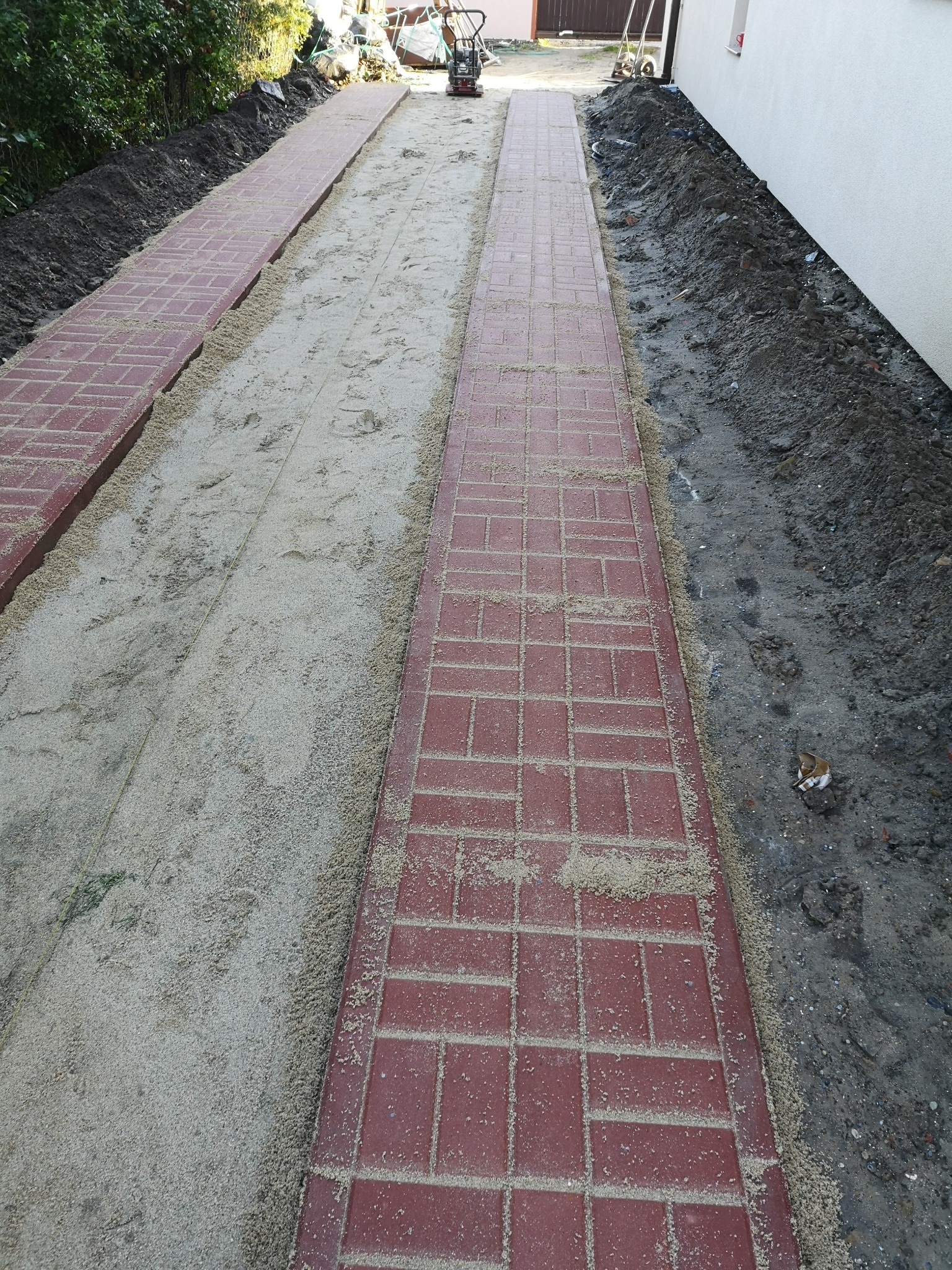 Czerwona kostka brukowa układana wzdłuż ścieżki, z piaskiem między kostkami i ubijarką w tle, widok z góry.