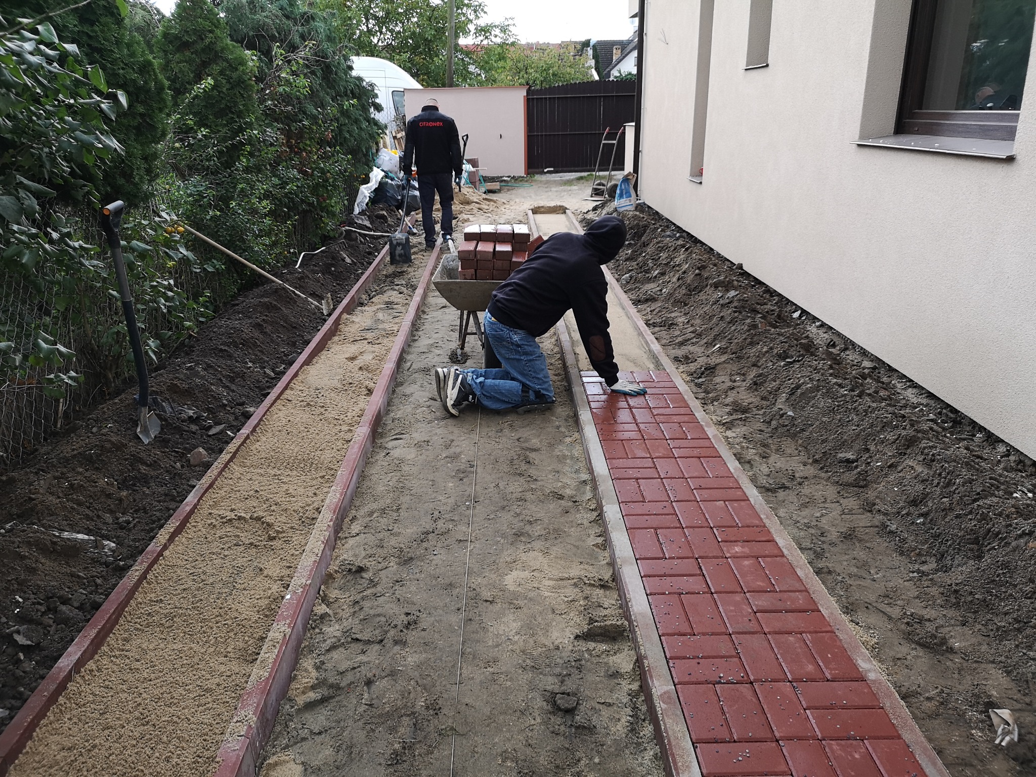 Układanie czerwonej kostki brukowej na ścieżce w ogrodzie, widoczny pracownik na kolanach, przygotowane podłoże z piasku, drugi pracownik w tle, dom z jasną elewacją.