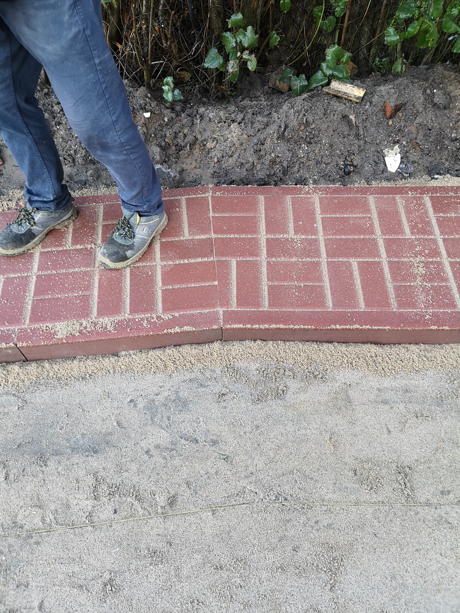 Układanie czerwonej kostki brukowej na podsypce piaskowej, widoczne buty robocze i fragment spodni osoby układającej, w tle ziemia i krzewy.