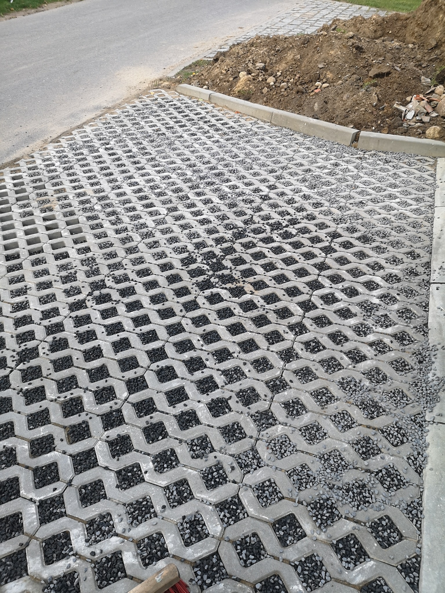 Szara, betonowa ażurowa nawierzchnia z wypełnieniem z drobnego, ciemnego grysu, widoczna krawędź chodnika i fragment drogi asfaltowej, w tle pryzma ziemi.
