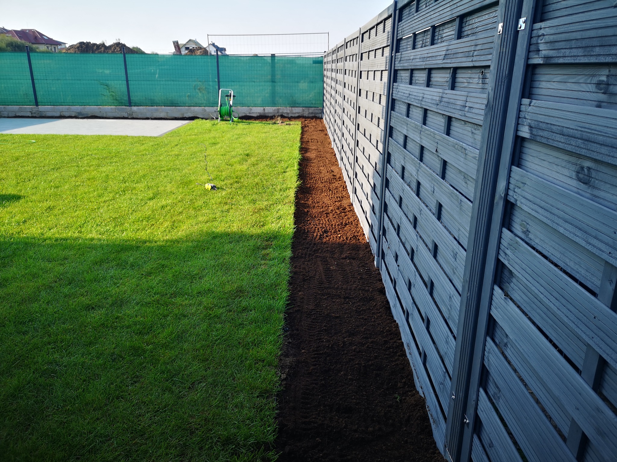 Szary, drewniany płot panelowy, świeżo przekopana ziemia wzdłuż ogrodzenia, trawnik, w tle siatka ogrodzeniowa i budowa.
