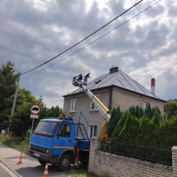 Przedsiębiorstwo Usługowe Friday - Dwóch pracowników na podnośniku koszowym czyści dach pokryty szarą blachą na tle pochmurnego nieba, widoczny fragment ulicy z zaparkowanym niebieskim samochodem i znak drogowy zakazu wjazdu...