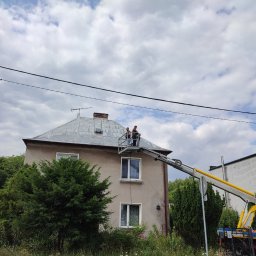 Przedsiębiorstwo Usługowe Friday - Dwaj pracownicy na podnośniku koszowym czyszczą metalowy dach dwupiętrowego domu z kominem, widok z poziomu gruntu, słup oświetleniowy po lewej stronie.