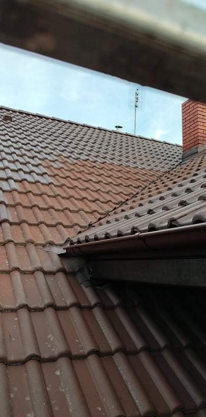 Częściowo oczyszczony, brązowy dach z dachówki ceramicznej, widoczna różnica w kolorze między czystą a zabrudzoną powierzchnią, rynna i komin w tle.