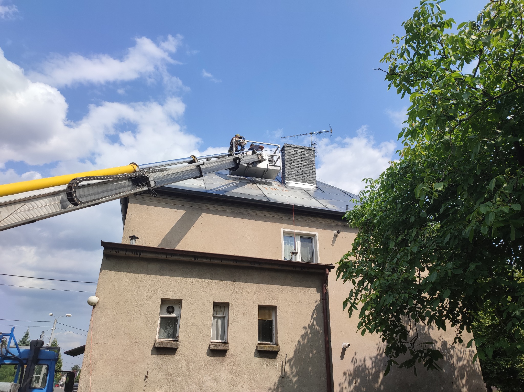 Pracownicy na podnośniku koszowym, czyszczący dach budynku z ceglanym kominem na tle błękitnego nieba z chmurami.