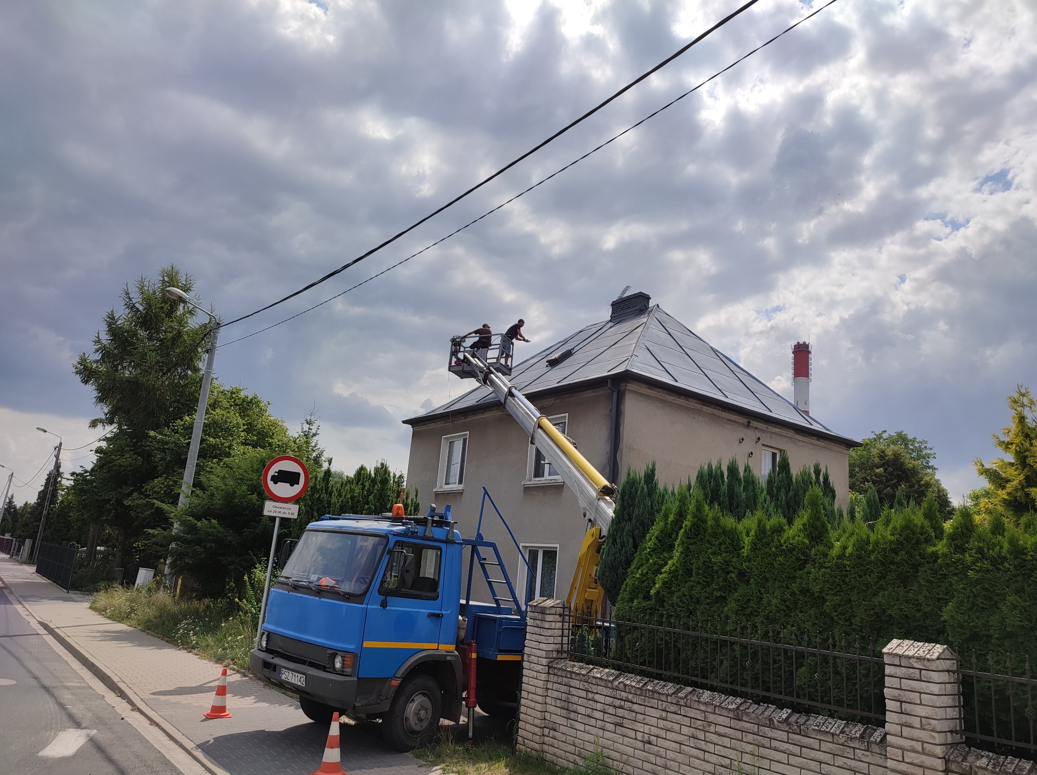 Dwóch pracowników na podnośniku koszowym czyści dach pokryty szarą blachą na tle pochmurnego nieba, widoczny fragment ulicy z zaparkowanym niebieskim samochodem i znak drogowy zakazu wjazdu...