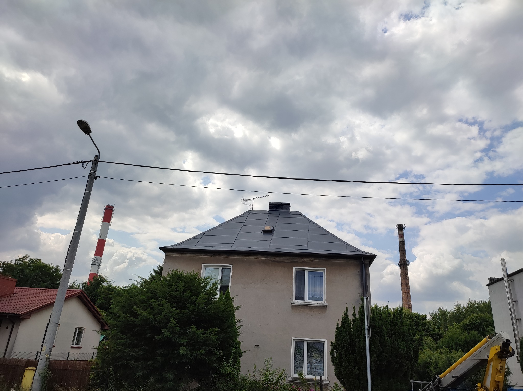 Dwupiętrowy dom z nowym, ciemnoszarym dachem na tle pochmurnego nieba, widoczny komin fabryczny w tle po lewej stronie, fragment podnośnika koszowego po prawej.