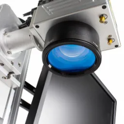 Zbliżenie na srebrną głowicę lasera z czarnym obiektywem F-Theta i niebieską soczewką, częściowo zasłaniającą ekran monitora.