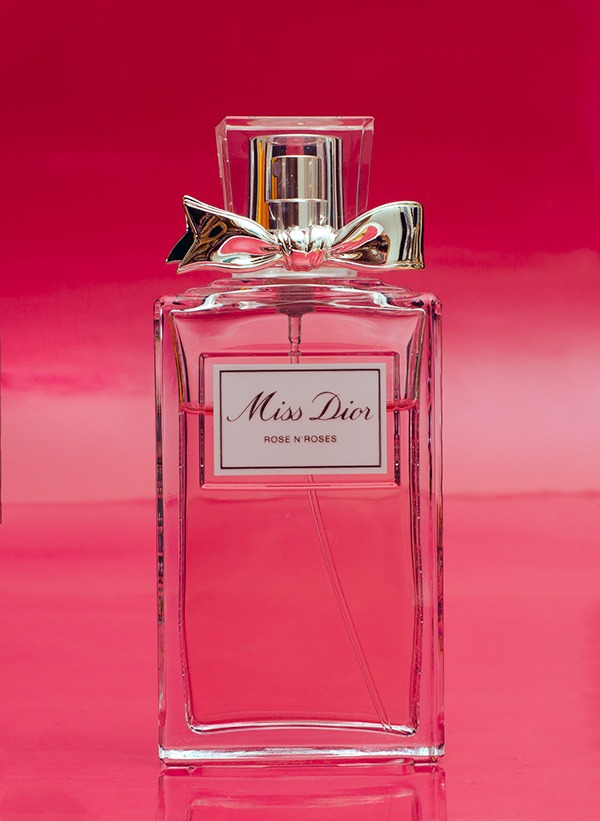 Butelka perfum Miss Dior Rose N'Roses na różowym tle z refleksami, srebrna kokarda na korku.