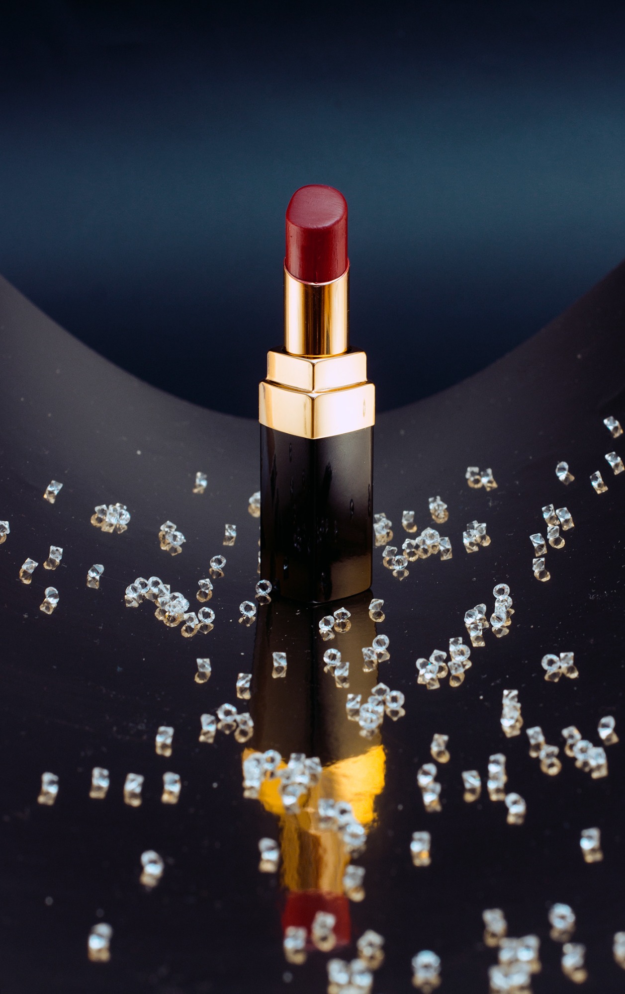 Szminka w czarno-złotym opakowaniu na błyszczącym tle z rozsypanymi, drobnymi, przezroczystymi kryształkami.