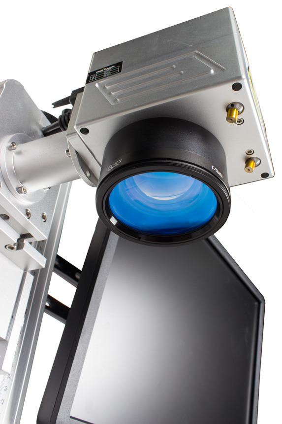 Zbliżenie na srebrną głowicę lasera z czarnym obiektywem F-Theta i niebieską soczewką, częściowo zasłaniającą ekran monitora.