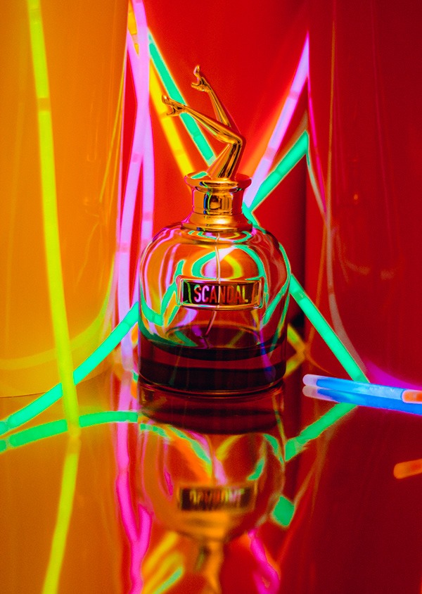 Flakon perfum Scandal z złotymi nogami na korku, otoczony neonowymi światłami w odcieniach różu, zieleni i pomarańczy, odbijający się w błyszczącej powierzchni.