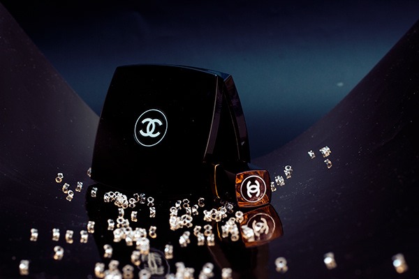 Dwa eleganckie opakowania kosmetyków z logo marki, otoczone drobnymi, błyszczącymi elementami na ciemnym, refleksyjnym tle.