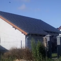 Nowy, ciemny dach z blachy na jasnym domu jednorodzinnym, widok z poziomu gruntu, w tle błękitne niebo i przelatujący ptak.