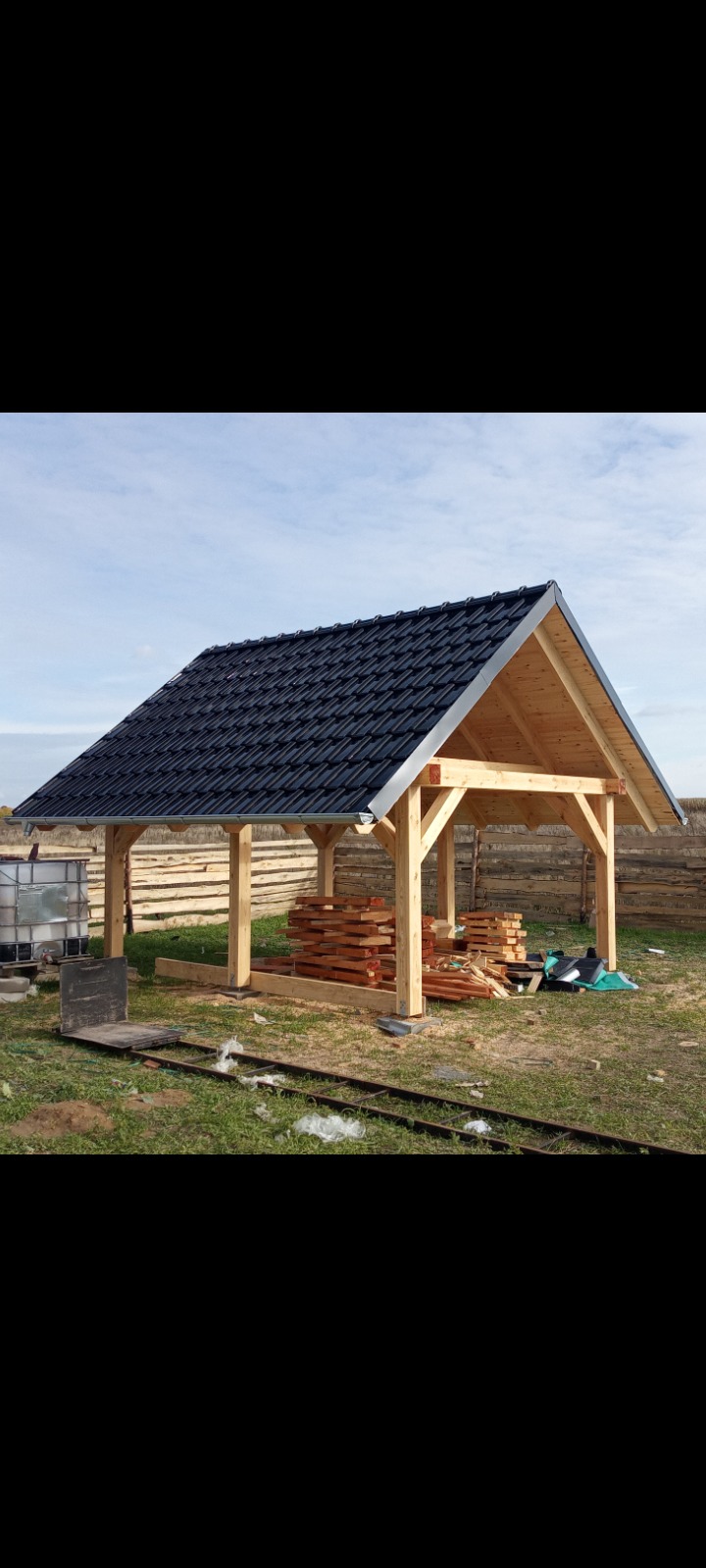 Drewniana altana z czarnym dachem z dachówki ceramicznej, widok z zewnątrz, w tle błękitne niebo. Częściowo składowane drewno wewnątrz konstrukcji.