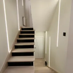 Nowoczesne schody z drewnianymi stopniami wykończonymi czarnym marmurem, oświetlenie LED wzdłuż ściany i pod stopniami, białe ściany i drzwi.