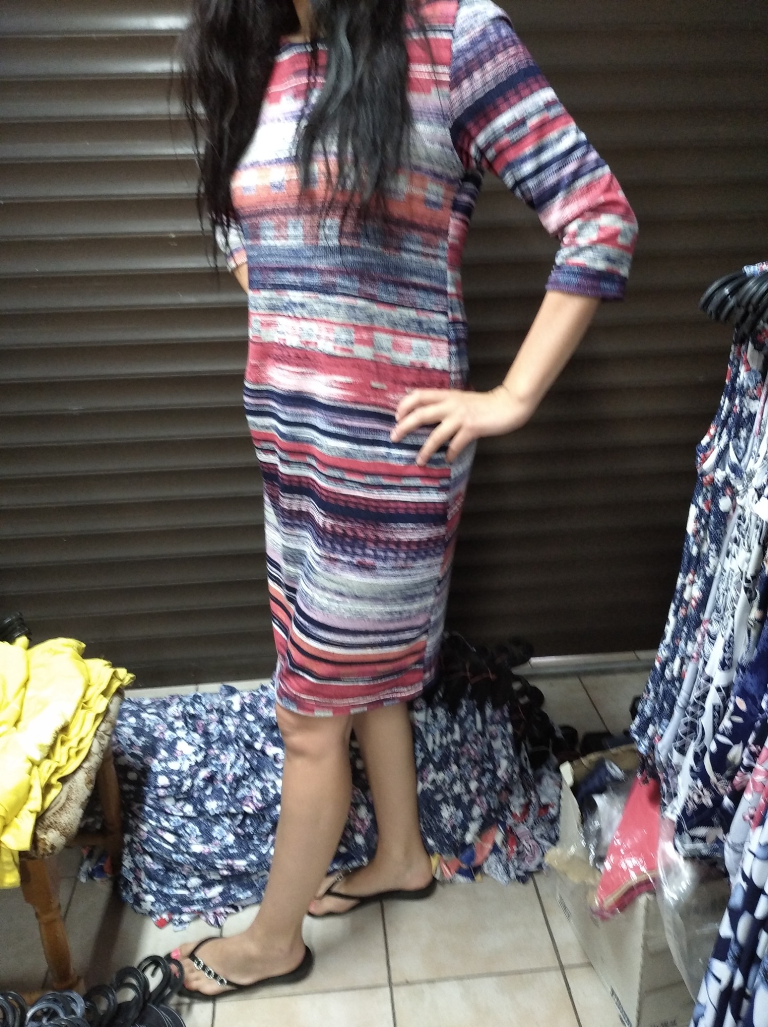 Kobieta prezentuje sukienkę w paski z rękawem 3/4, stojąc na tle metalowej rolety i sterty ubrań.