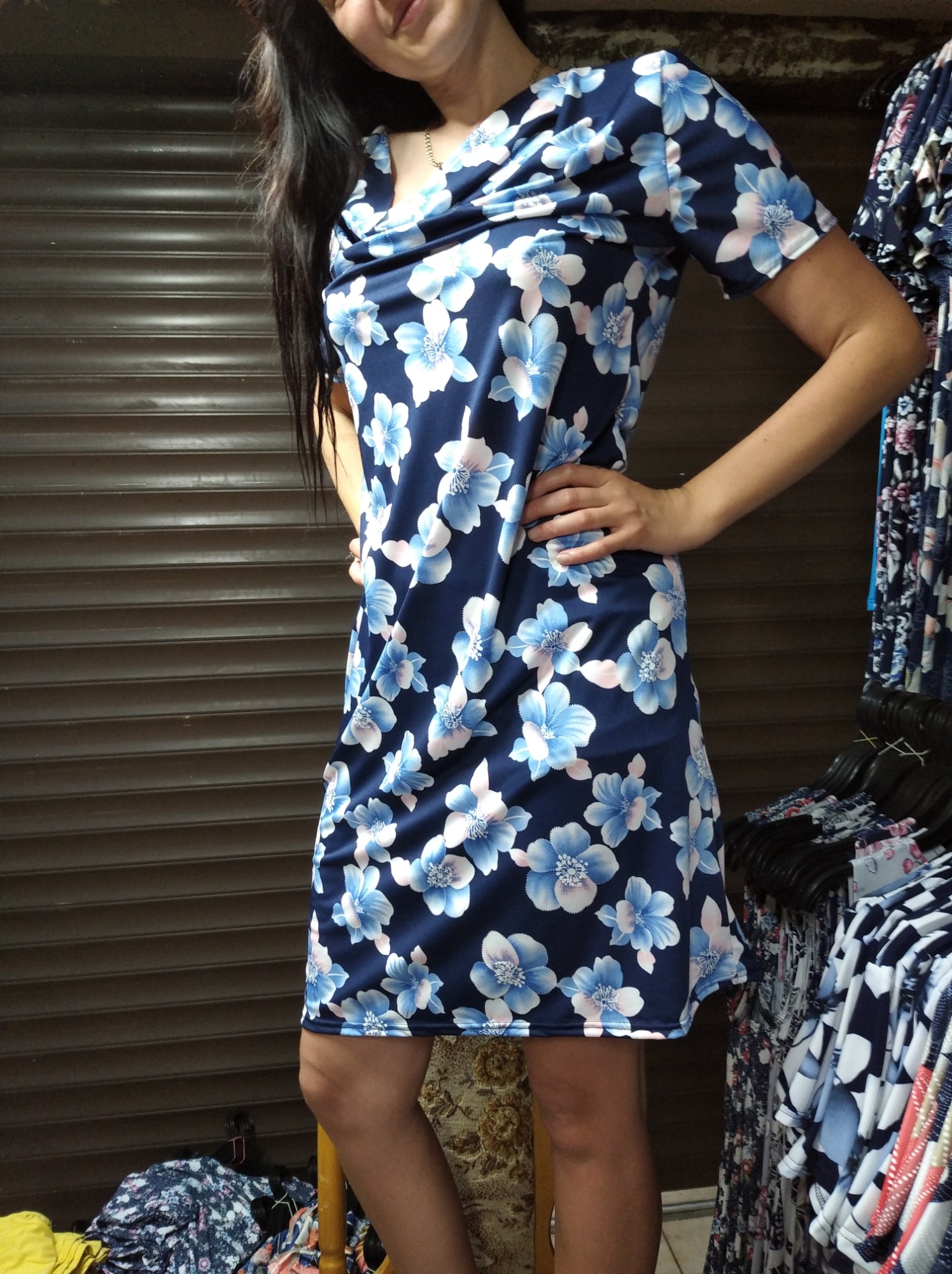 Kobieta prezentuje letnią sukienkę w kwiaty na tle metalowej rolety i wieszaków z odzieżą.