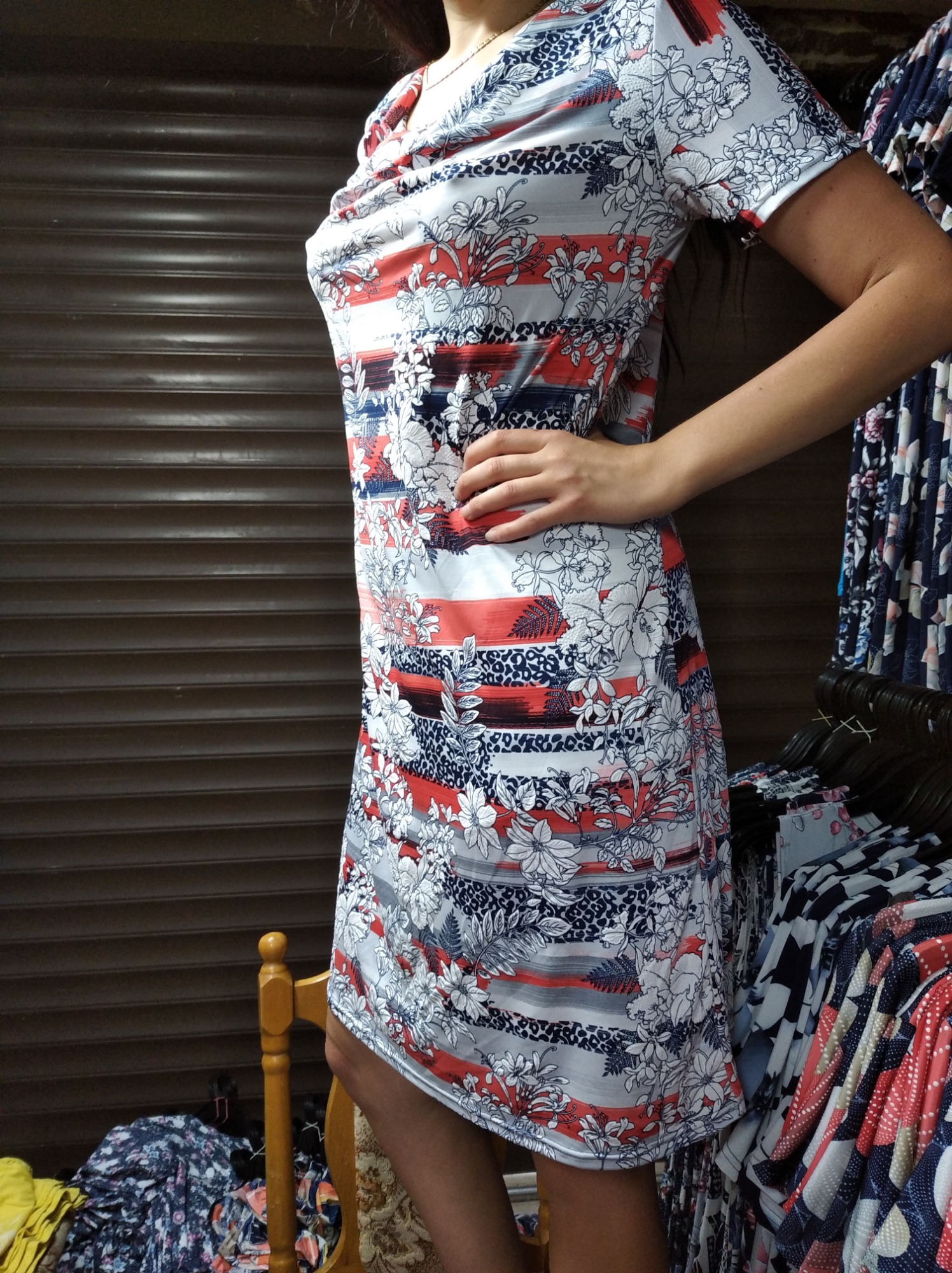 Krótka sukienka damska z wzorem kwiatowym i geometrycznym, prezentowana na osobie w szwalni odzieży, z widocznymi w tle wieszakami z innymi ubraniami i zamkniętą roletą.