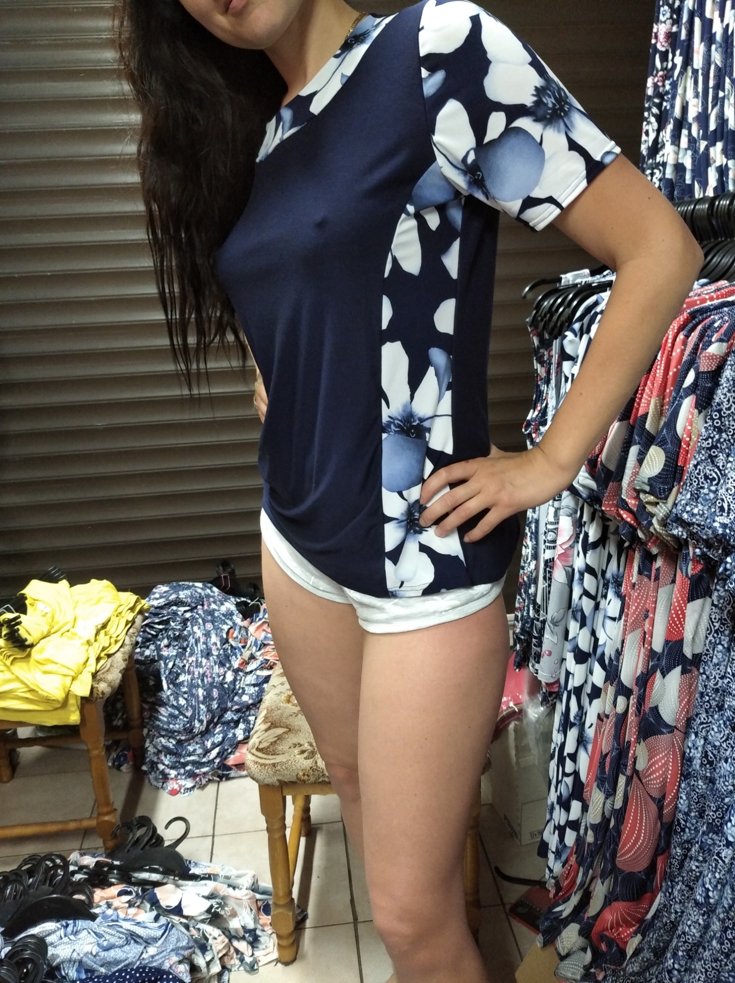 Kobieta prezentuje granatową bluzkę z białymi kwiatami na ramionach, stojąc w pomieszczeniu z wieszakami pełnymi ubrań z wzorzystych materiałów.