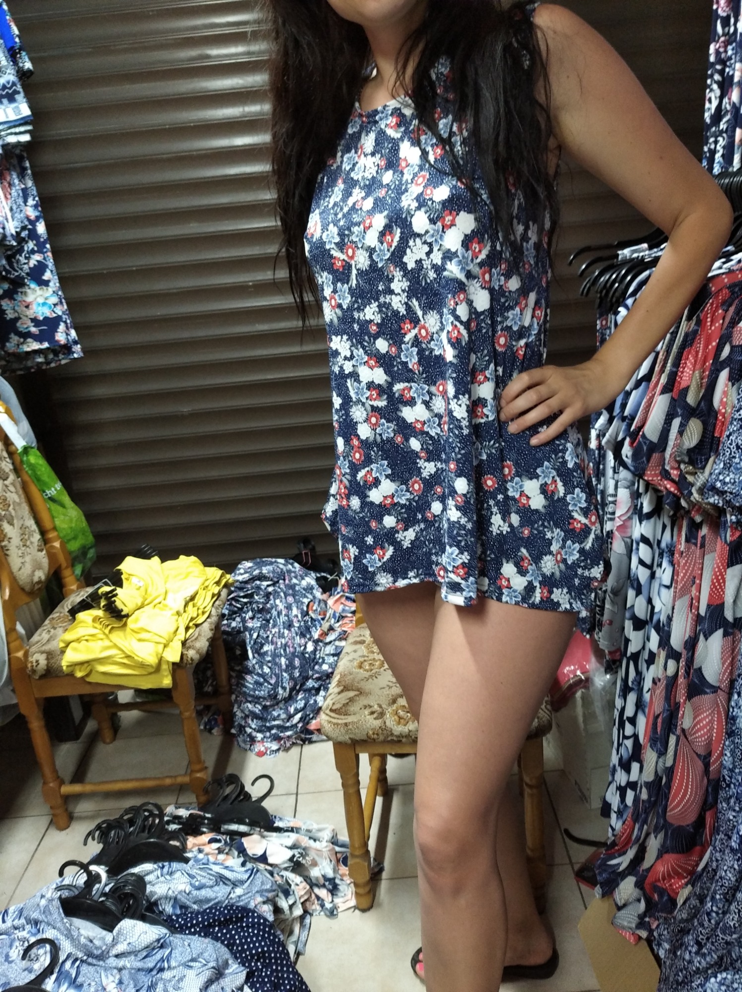 Kobieta prezentuje letnią sukienkę w kwiatowy wzór na tle wieszaków z ubraniami i sterty odzieży na krzesłach.