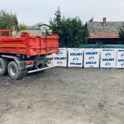 Czerwona ciężarówka z wywrotką zaparkowana obok palet z materiałem budowlanym Solbet na placu budowy. W tle widoczny dom z ceglanym kominem i drzewa.