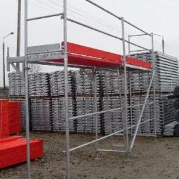 Srebrne, metalowe rusztowanie z czerwonymi elementami na tle stosów blachy trapezowej i czerwonych desek, widoczne na zewnątrz w pochmurny dzień.