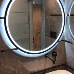 Nowoczesna łazienka z okrągłym lustrem LED, umywalką nablatową i armaturą w kolorze matowej czerni na tle płytek imitujących marmur z delikatnymi żyłami.