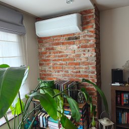 APJ-System - Biała klimatyzacja Samsung WindFree zamontowana na ceglanej ścianie w pokoju z rośliną, książkami i lampami. Widok na okno z roletami.