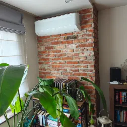 Biała klimatyzacja Samsung WindFree zamontowana na ceglanej ścianie w pokoju z rośliną, książkami i lampami. Widok na okno z roletami.